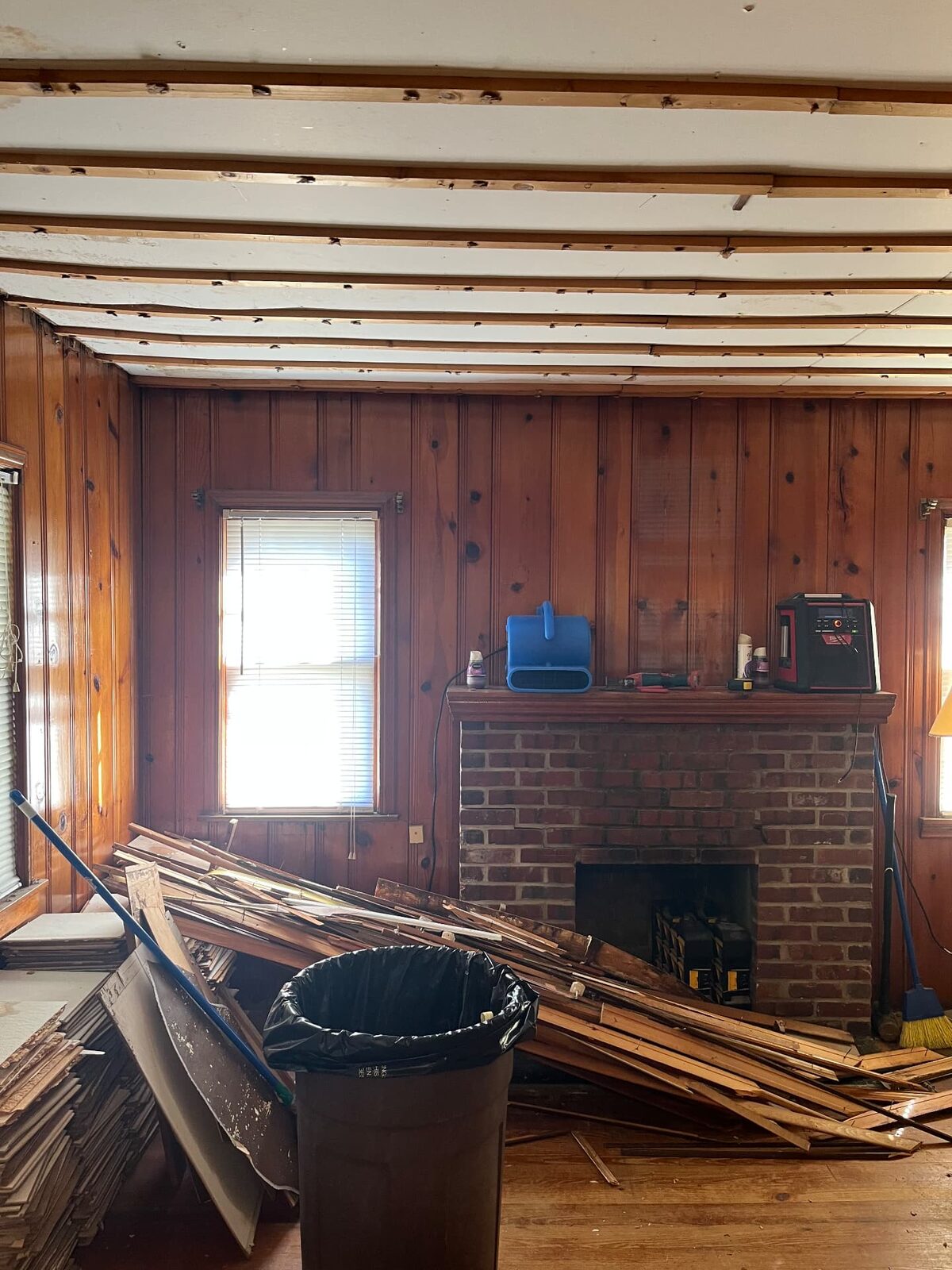 Room with wood paneling and ceiling material being torn out, debris stacked on the floor