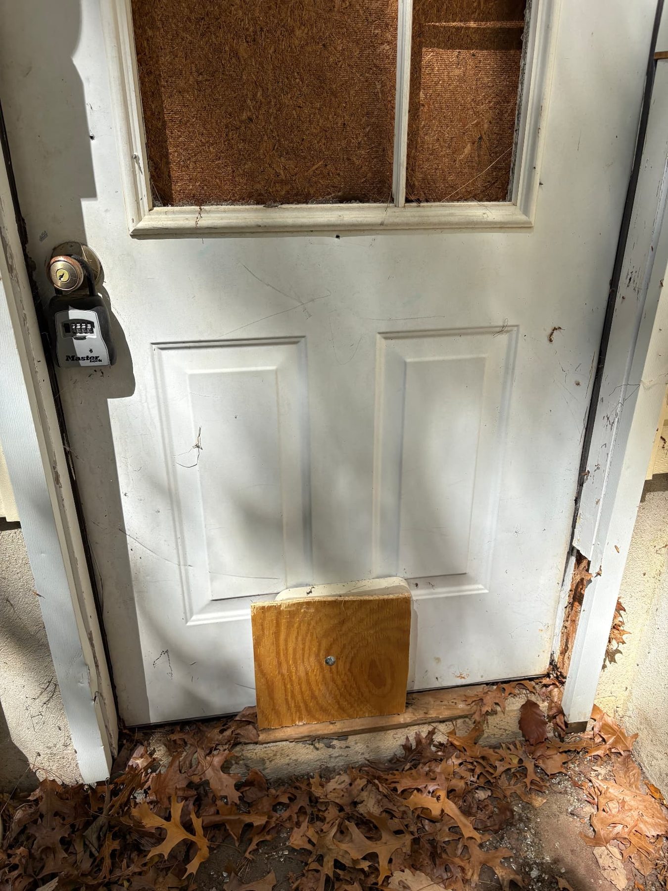 Vacant property entry door secured with a combination lockbox, a boarded glass panel and a wood repair block at the base