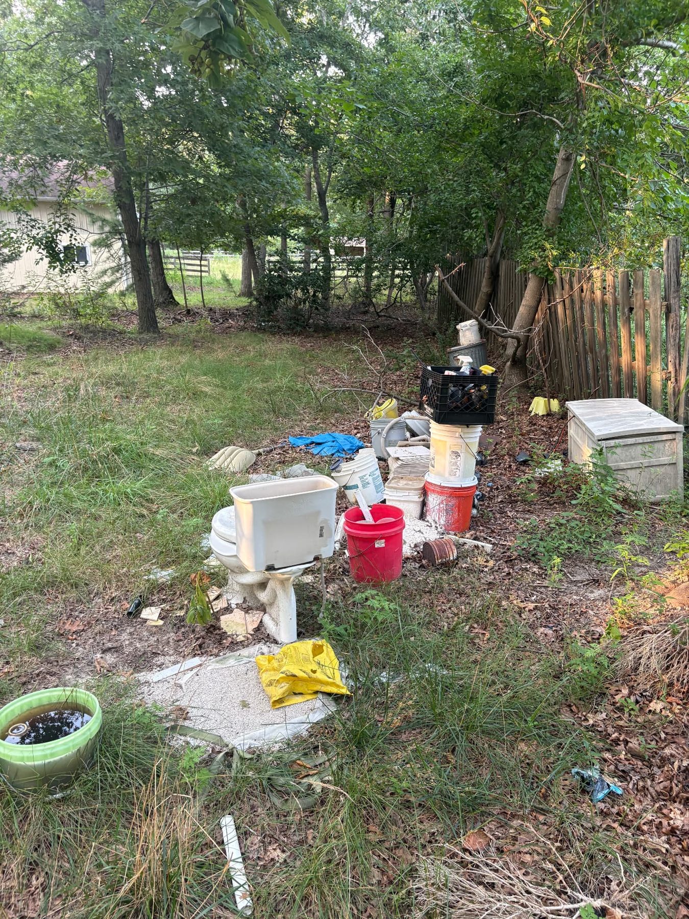Rear yard of a vacant property with a discarded toilet, buckets, crates and scattered debris along the fence line